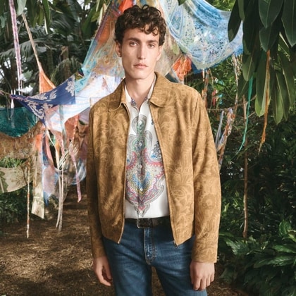 Model in a gold paisley jacket over a colorful patterned Etro shirt, standing in a lush garden with draped scarves behind him.