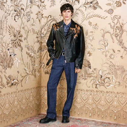 Model wearing a dark leather jacket with floral embroidery over a patterned shirt and blue trousers.