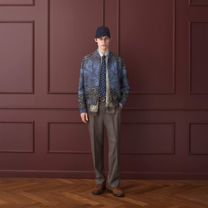 Model wearing an Etro patterned bomber jacket layered over a shirt and tie, paired with tailored trousers, brown shoes, and a navy baseball cap