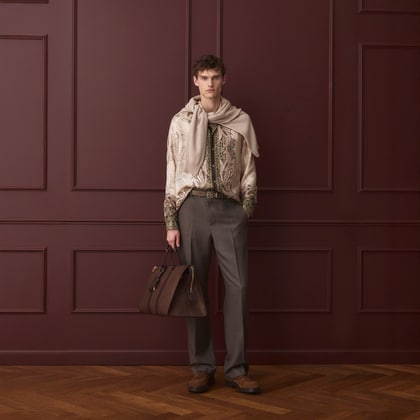 Model wearing a paisley-print shirt with a sweater draped over the shoulders, tailored trousers, and carrying a leather bag.