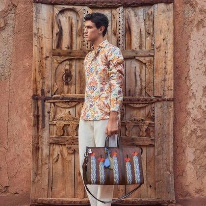 Model wearing a floral Etro shirt and cream trousers, holding a brown leather bag with colorful embroidered motifs, standing in front of a rustic wooden door.