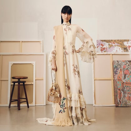 Model wearing a flowing floral Etro gown with ruffled sleeves and hem, styled with an embroidered scarf and tasseled bag.