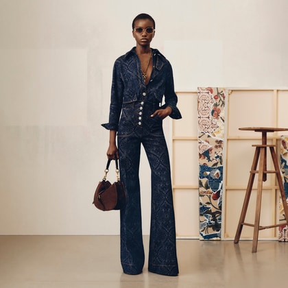 Model wearing a dark blue patterned jacket and flared pants, holding a brown handbag, standing near an easel with colorful artwork. 