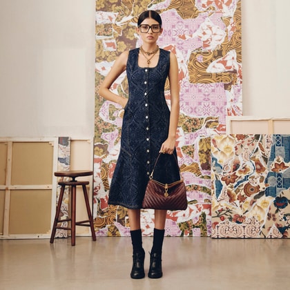 Model wearing a sleeveless button-down Etro dress with a paisley pattern, black ankle boots, and holding a quilted bag, standing against an artistic collage background.