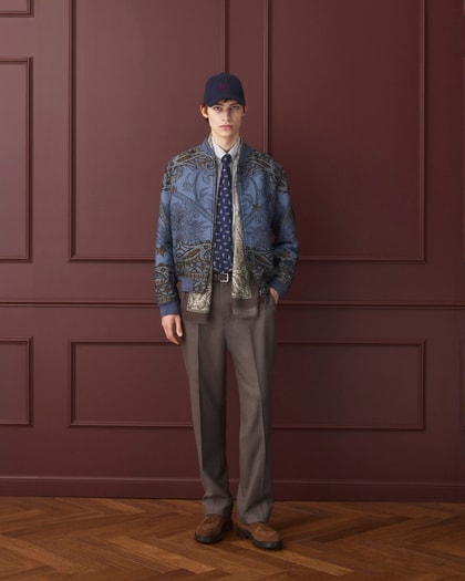 Model wearing an Etro patterned bomber jacket layered over a shirt and tie, paired with tailored trousers, brown shoes, and a navy baseball cap