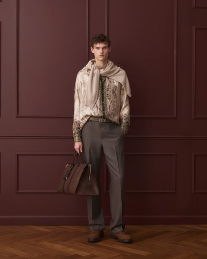 Model wearing a paisley-print shirt with a sweater draped over the shoulders, tailored trousers, and carrying a leather bag.