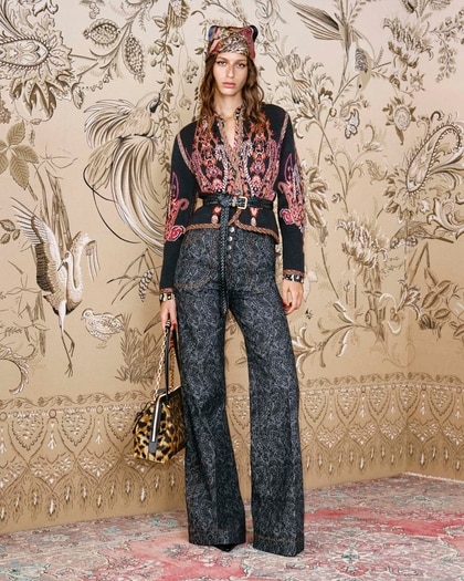 Model wearing an eclectic Etro outfit featuring a patterned black and red jacket cinched with a braided belt, wide-leg printed trousers, a leopard-print bag, and a coordinating headscarf, standing against a detailed bird-and-floral wallpaper backdrop.