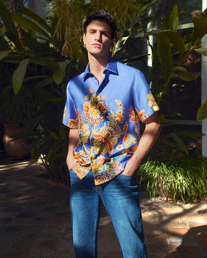 Male model wearing a vibrant blue, floral Etro shirt paired with jeans, standing in a lush, sunlit garden setting.