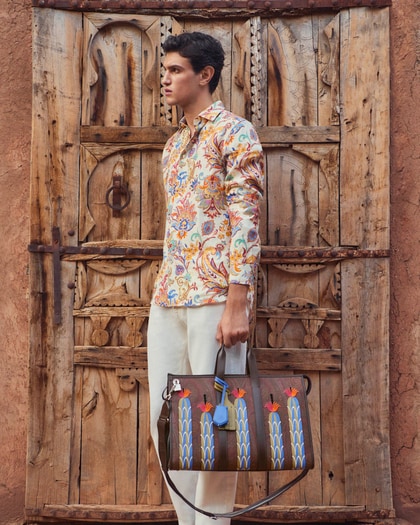 Model wearing a floral Etro shirt and cream trousers, holding a brown leather bag with colorful embroidered motifs, standing in front of a rustic wooden door.