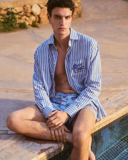 Model wearing an Etro striped button-down shirt paired with printed swim shorts, seated poolside in a relaxed summer setting.
