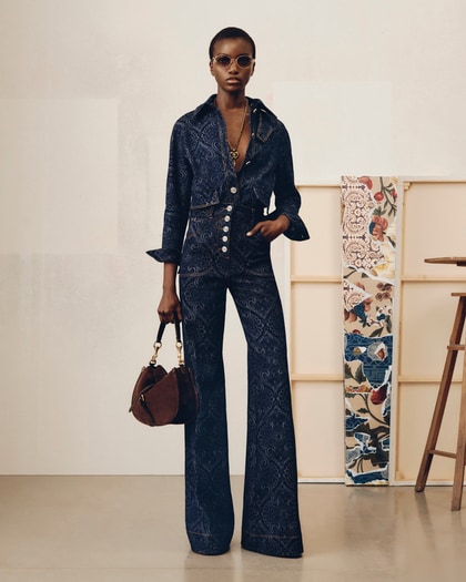 Model wearing a dark patterned Etro jumpsuit with flared trousers, accessorized with sunglasses, a necklace, and a brown handbag.