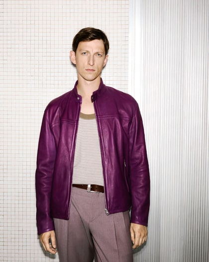 The model wears a purple leather jacket over a striped tank, paired with taupe trousers and a brown belt