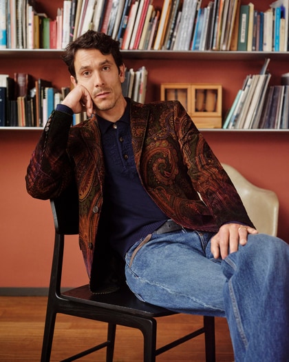 Man wearing a paisley jacket and navy polo shirt, seated in front of bookshelves.