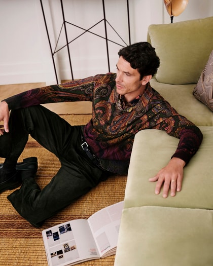Man wearing an ETRO paisley shirt and black trousers, relaxing on a sofa.