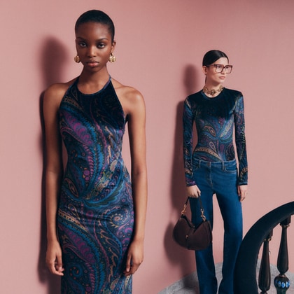 Two models wearing ETRO outfits with a vibrant paisley print. The front model, standing against a soft pink background, wears a sleeveless halter-neck dress with shades of blue and pink.