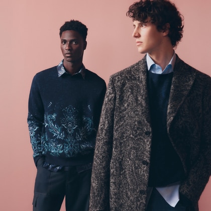 Two individuals standing against a light pink background, wearing patterned sweaters and coats with faces obscured.