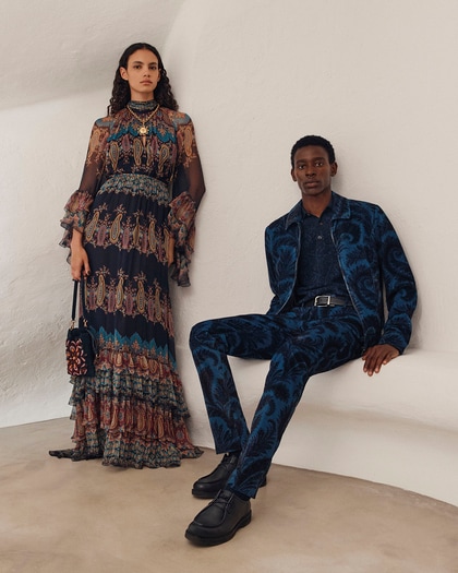 Two models wearing Etro outfits — the woman in a long patterned bohemian dress and the man in a blue patterned suit.