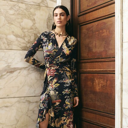 Model wearing a long floral print wrap dress with a front slit, standing against a marble wall.