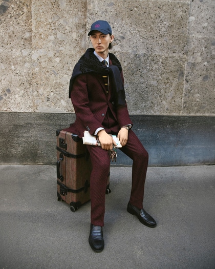 Model wearing an Etro burgundy tailored suit with a navy cape, white shirt, tie, and baseball cap, seated on a vintage suitcase against a stone wall.