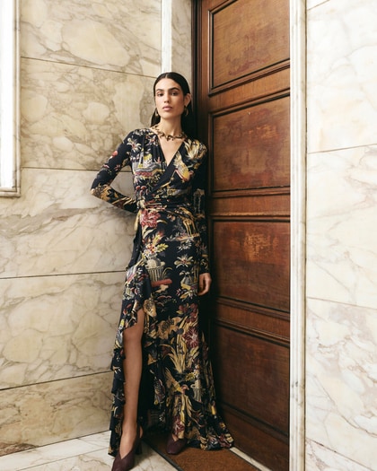 Model wearing a long floral print wrap dress with a front slit, standing against a marble wall.