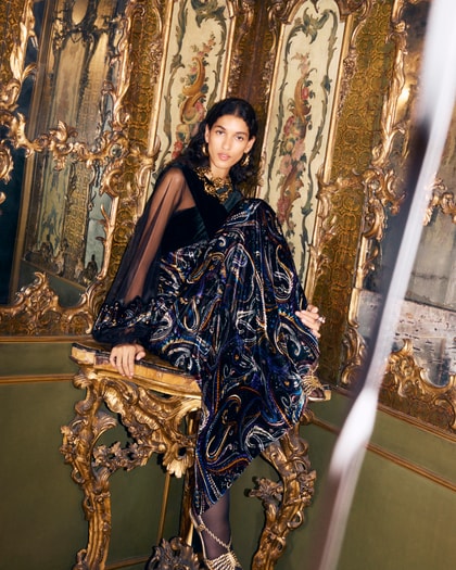 Model wearing an elegant Etro gown with a dark paisley pattern and sheer sleeves, seated on an ornate gilded chair in a richly decorated room.