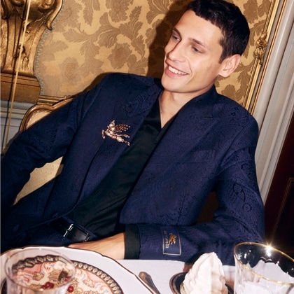 Model wearing a navy patterned Etro jacket with a decorative brooch, seated at an elegant dining table