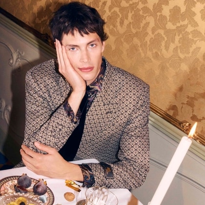 Model in an ornate patterned Etro jacket seated at an elegant table set with fruit, sweets, and candlelight.