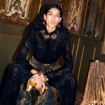 Model in an ornate black velvet Etro outfit with gold accessories, seated in an opulent, richly decorated room.