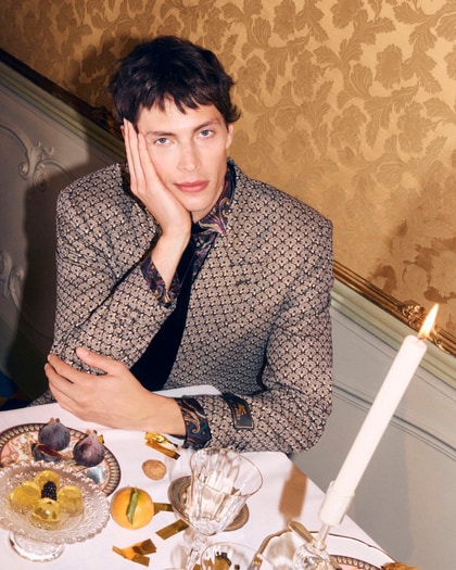 Model in an ornate patterned Etro jacket seated at an elegant table set with fruit, sweets, and candlelight.