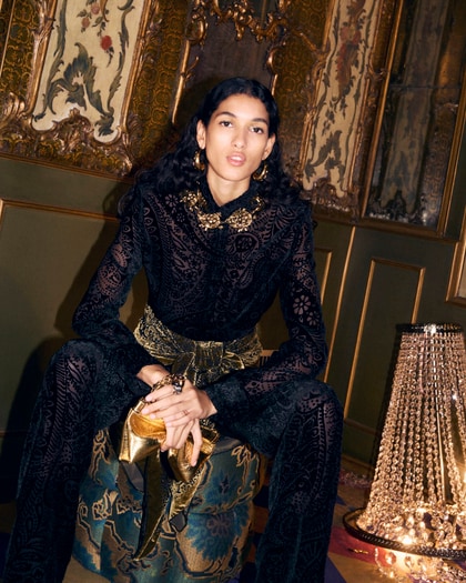Model in an ornate black velvet Etro outfit with gold accessories, seated in an opulent, richly decorated room.