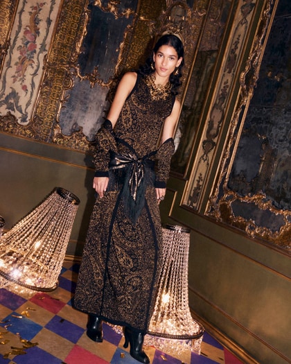Model wearing a long black and gold Etro dress with ornate patterns, posed in an opulent room with decorative chandeliers