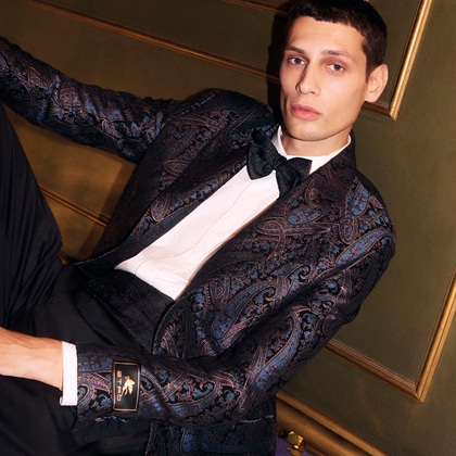 Model wearing an Etro tuxedo with a dark paisley-patterned jacket, white shirt, and black bow tie, seated against a gold-toned background
