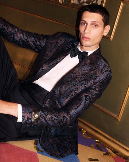 Model wearing an Etro tuxedo with a dark paisley-patterned jacket, white shirt, and black bow tie, seated against a gold-toned background