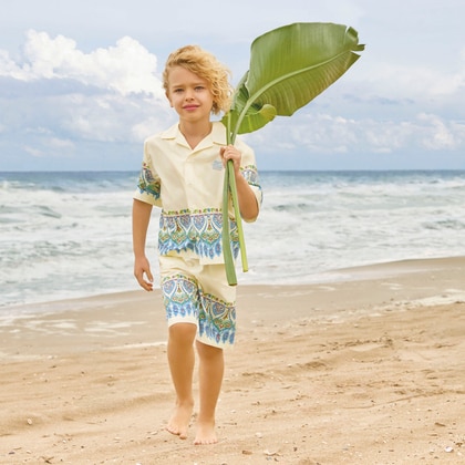 Young boy walking barefoot on the beach wearing an Etro printed outfit and holding a large green leaf by the sea.