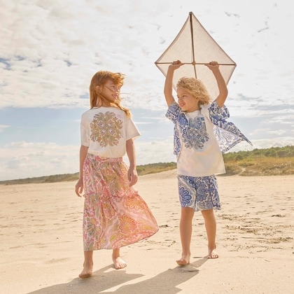 Two children walk barefoot along a sandy beach under a lightly clouded sky, dressed in colorful Etro outfits: the girl wears a white T-shirt with an ornate paisley motif and a flowing patterned skirt, while the boy wears a white and blue paisley-print set and holds a kite above his head.