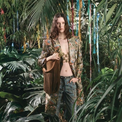Model in a lush jungle setting wearing a paisley Etro coat and matching trousers, layered over a jeweled top with a brown fringe bag.