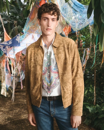 Model in a gold paisley jacket over a colorful patterned Etro shirt, standing in a lush garden with draped scarves behind him.