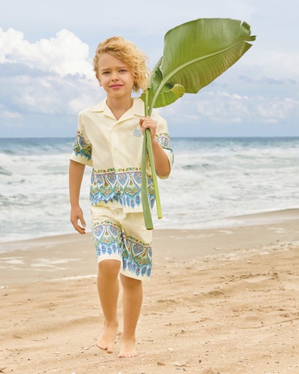 Young boy walking barefoot on the beach wearing an Etro printed outfit and holding a large green leaf by the sea.