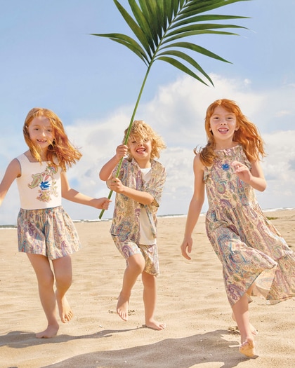 Three children running barefoot on a sandy beach, smiling and wearing Etro outfits, one holding a large palm leaf under a sunny sky.