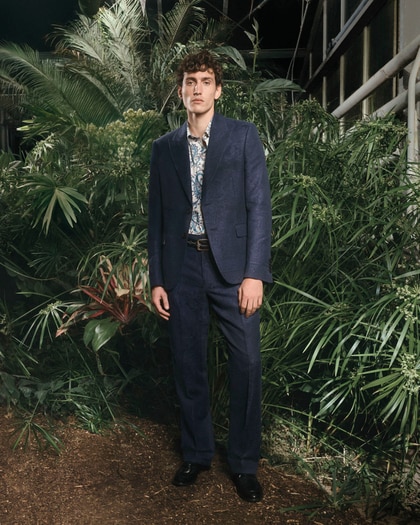 Male model in a navy patterned suit and paisley shirt, standing in lush greenery, wearing Etro.