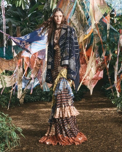 Model in a layered bohemian Etro gown and patterned cape walking through lush greenery with colorful scarves overhead.