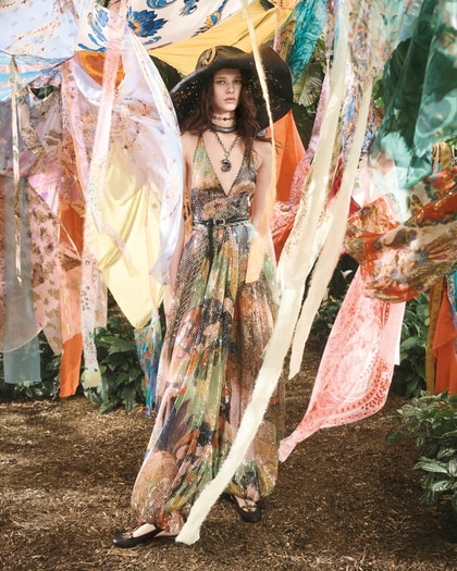 Model in a flowing printed maxi dress and wide-brim hat standing among hanging pastel fabrics in a garden setting.