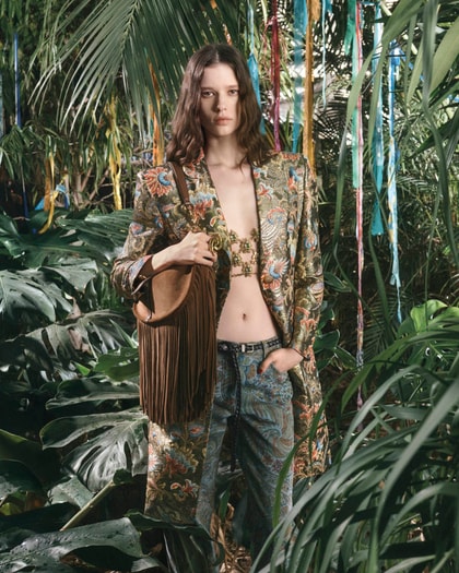 Model in a lush jungle setting wearing a paisley Etro coat and matching trousers, layered over a jeweled top with a brown fringe bag.