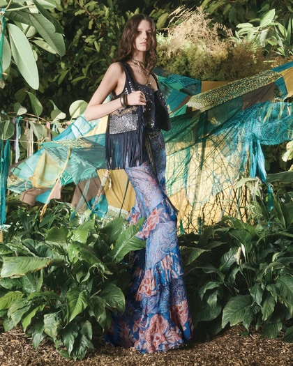 Model in a paisley skirt and fringed top posing in tropical foliage with draped scarves behind her.