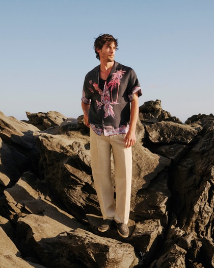 Man wearing an Etro printed short-sleeve shirt and relaxed trousers, standing on coastal rocks.