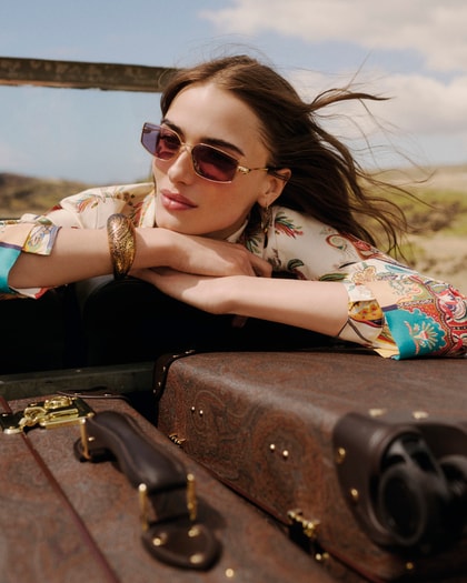 Woman wearing an Etro paisley-print shirt and matching scarf, accessorised with sunglasses and jewellery, leaning on vintage luggage outdoors.