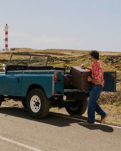 Man wearing an ETRO printed shirt with jeans beside a vintage off-road vehicle in a desert landscape.