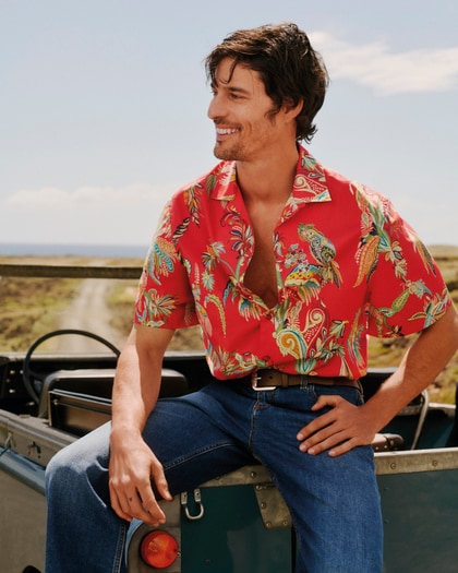 Model wearing an Etro red short-sleeve paisley-print shirt with blue jeans, sitting on the back of a vintage off-road vehicle