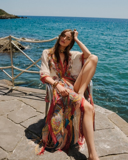 Woman wearing a flowing Etro paisley beach cover-up, seated by the seaside on a sunny day.