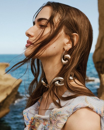 Side profile of a model by the sea wearing Etro, with windblown hair and sculptural silver swirl earrings and matching necklace.